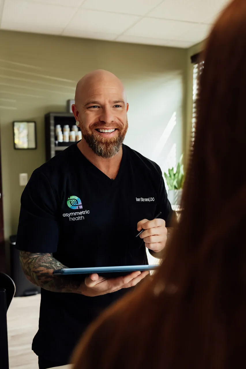 Dr. Ian Strand talking with a patient and reviewing notes on a tablet during a 60-90 minute evaluation at Asymmetric Health
