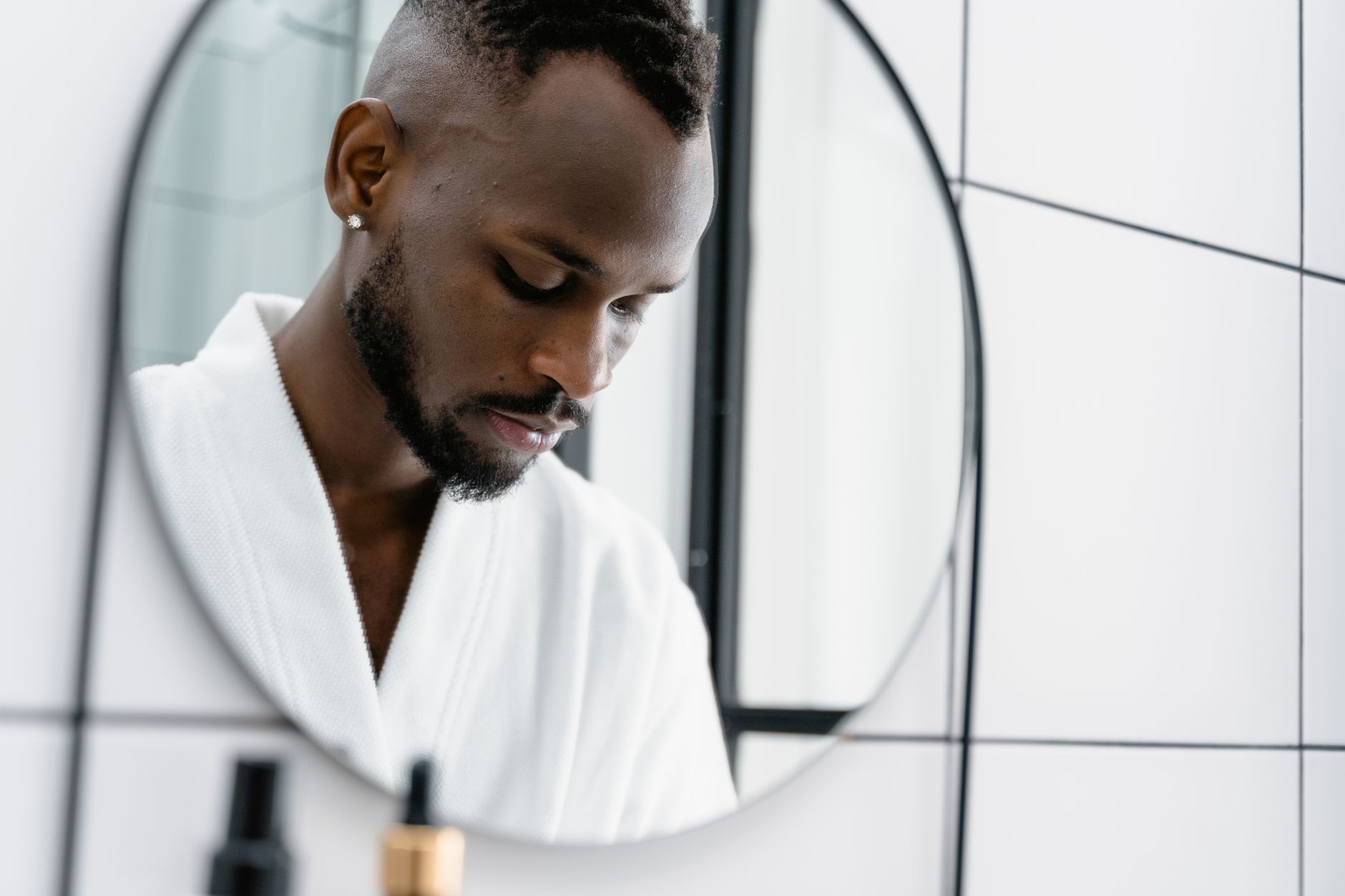 Thoughtful man looking down at his reflection in a modern bathroom mirror, illustrating an educational article on testosterone therapy and male pattern hair loss