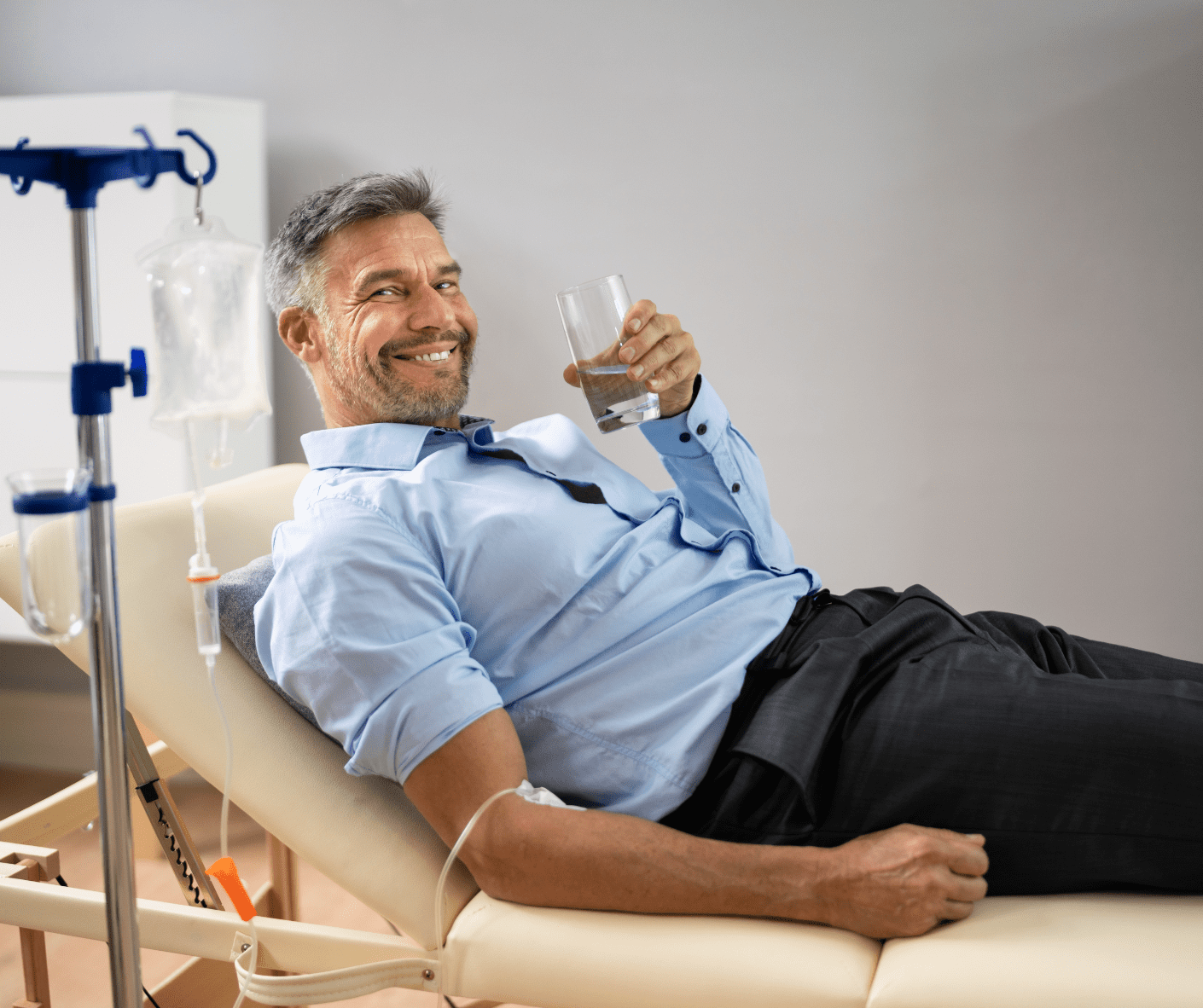 A man with an IV in his arm is sitting, smiling, and holding a glass of water
